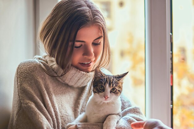 Bella ragazza in un maglione caldo abbraccia il suo gatto preferito seduto sul davanzale della finestra accanto alla finestra aperta