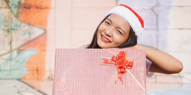A beautiful girl wear Christmas hat hold present on colorful background.
