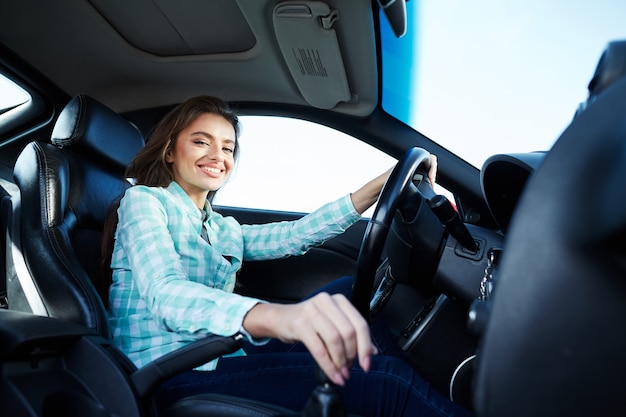 Bella ragazza che indossa la camicia blu seduto nella nuova automobile, felice, bloccato nel traffico, ascoltando la musica, ritratto, guardando la fotocamera e sorridente.