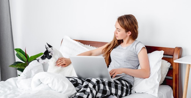 ノートパソコンとベッドに座ってかわいい犬をかわいがってパジャマを着ている美しい少女。ノートブックに取り組んでいる朝の時間にペットと一緒に家で若い女性。