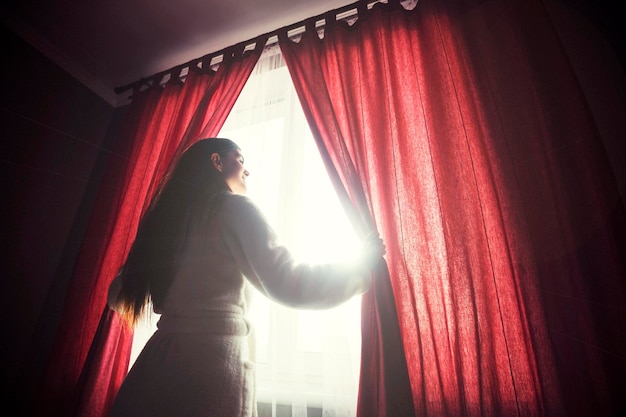 Beautiful girl wears white bathrobe opening curtains on the morning window with sunrise