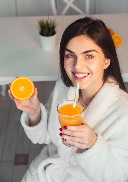 Beautiful girl wears white bathrobe with glass of orange juice and fruit kitchen morning sunset view