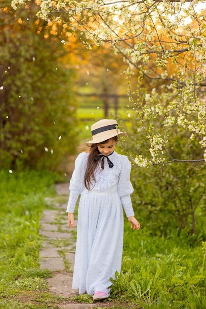 A beautiful girl in a white dress and a hat walks under flowering trees in the spring in the garden