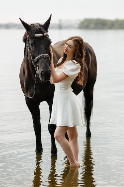 A beautiful girl in a white dress leads a horse across the river