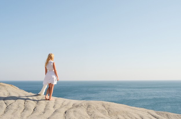 Bella ragazza in abito bianco in piedi su una roccia e guardando il mare