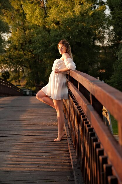 A beautiful girl in a white dress stands on a wooden bridge