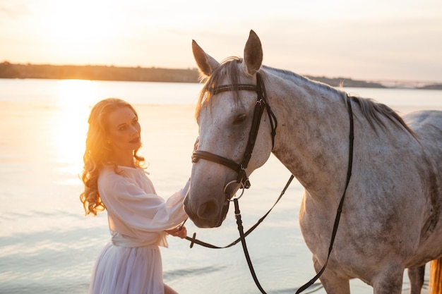 Beautiful girl in a white dress and a white horse on the beach against the backdrop of sunset