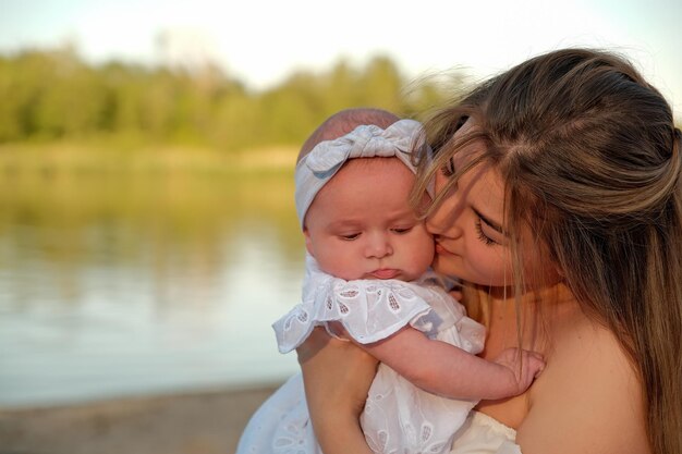 Photo a beautiful girl in a white dress with a newborn baby is sitting on the beach on the sand