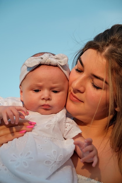 A beautiful girl in a white dress with a newborn baby is sitting on the beach on the sand