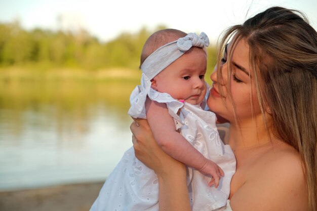 A beautiful girl in a white dress with a newborn baby is sitting on the beach on the sand