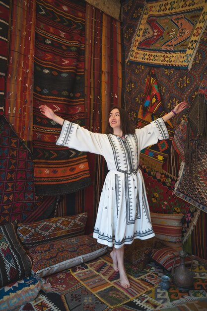 Beautiful girl in a white dress with a teapot in a traditional turkish interior with many carpets on the wall