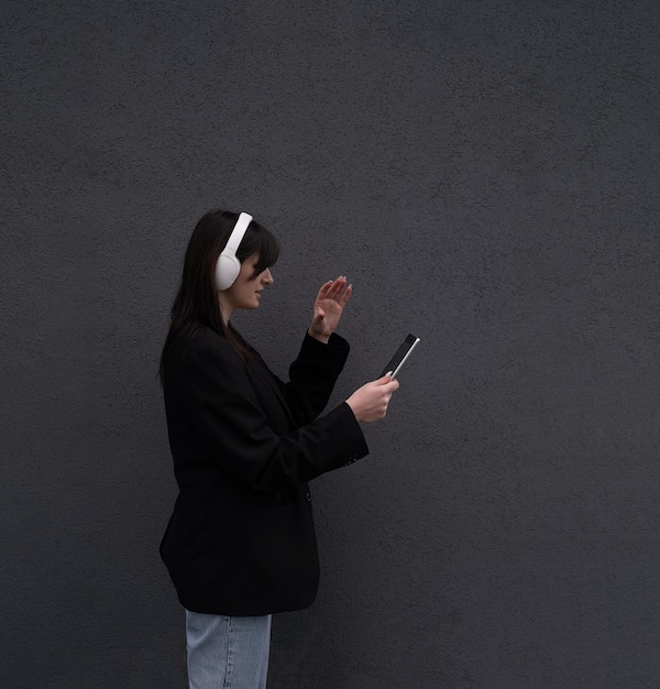 A beautiful girl in white headphones and a black jacket holds a tablet outside work outside listen