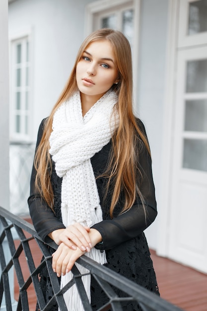 Beautiful girl in a white knitted scarf and a black fashionable dress standing on the veranda of house