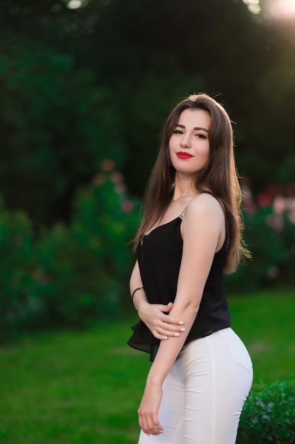 A beautiful girl in white pants and a black sweater posing against a background of flowers