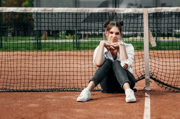 A beautiful girl in a white shirt, black pants and white sneakers sits at the net on the tennis court.