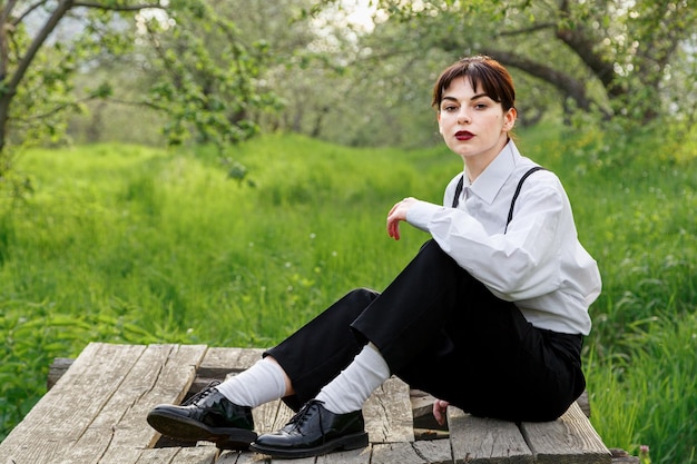 A beautiful girl in a white shirt in black trousers with suspenders against the background of the sky and green grass close up