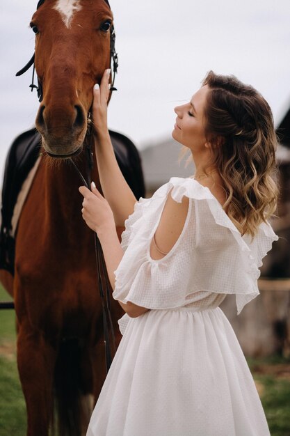 古い牧場の馬の横にある白いサンドレスの美しい少女
