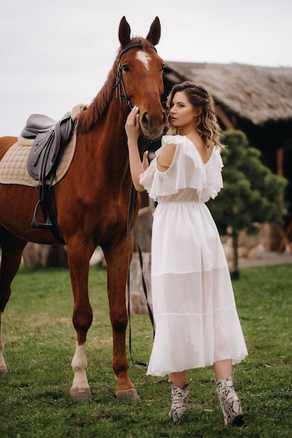 Красивая девушка в белом сарафане рядом с лошадью на старом ранчо