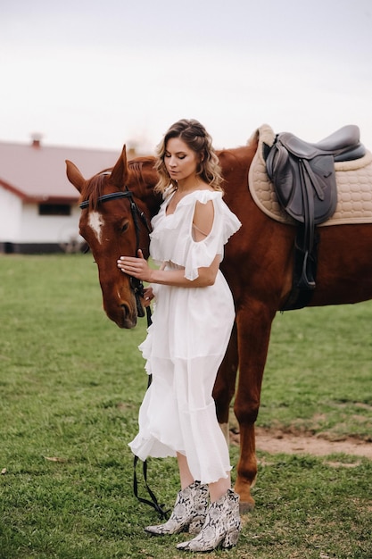 Beautiful girl in a white sundress next to a horse on an old ranch