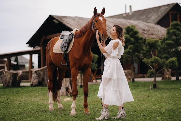 Beautiful girl in a white sundress next to a horse on an old ranch