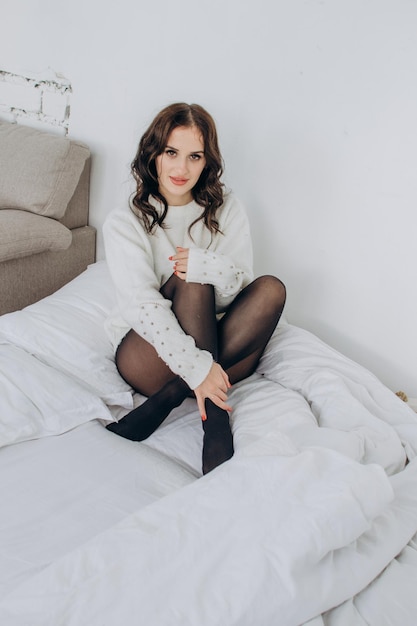 Beautiful girl in a white sweater and black tights sits on the bed