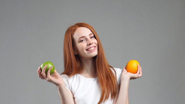 Bella ragazza in una maglietta bianca sorridente che tiene una mela in una mano e un pompelmo nell'altra, su uno sfondo grigio. cibo sano, frutti deliziosi