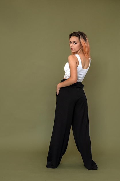 Beautiful girl in a white top and black pants stands beautifully on a dark green background