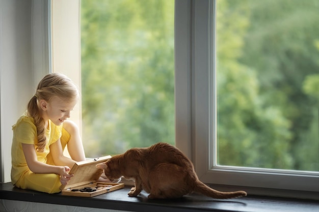 Beautiful girl on the windowsill plays checkers with a cat