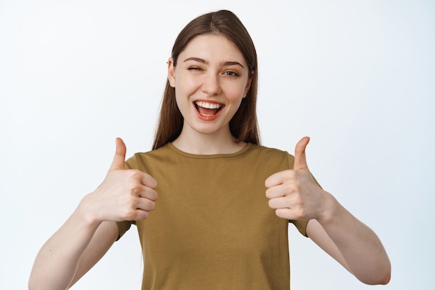 Beautiful girl winking and smiling, showing thumbs up to compliment product, praise quality, assuring you, standing on white.