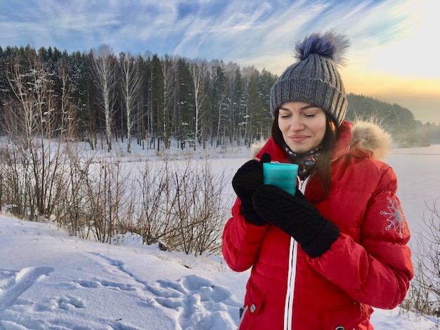 Beautiful girl in a winter jacket with a mug in winter in nature. A girl in a hat and a red warm jacket. He warms his hands and drinks hot tea or a drink