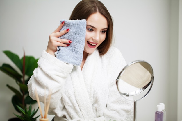 Beautiful girl wipes her face with a towel at home in front of a mirror