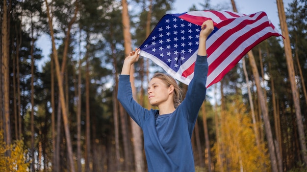 アメリカの国旗を持つ美しい少女。暖かい夏の森の手にアメリカの国旗