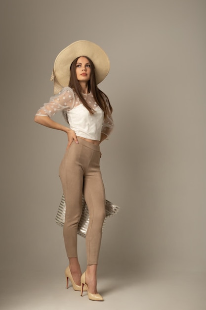 Beautiful girl with beach bag and hat poses on a white cyclorama. Beautiful woman with a model appearance on a light background