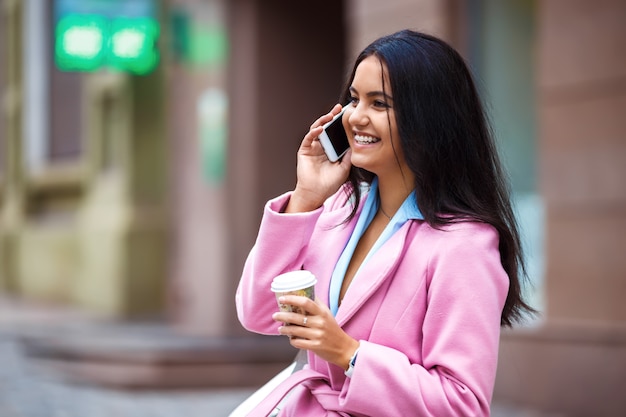 A beautiful girl with a beautiful smile talking on the phone. Young beautiful pretty girl walking along the street with handbag and cup of coffee and phone in her hands