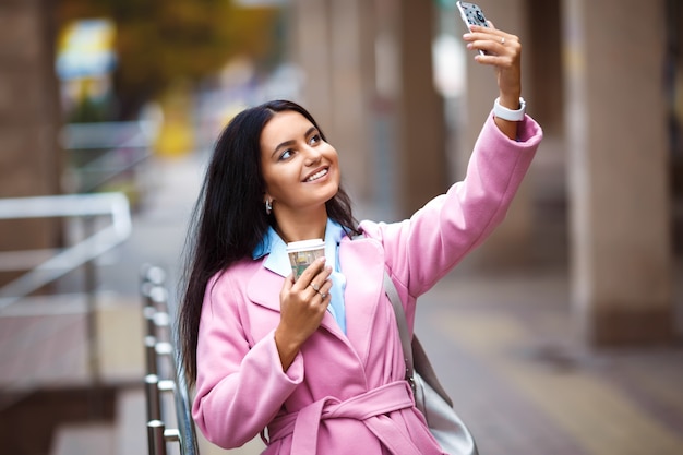 A beautiful girl with a beautiful smile talking on the phone. Young beautiful pretty girl walking along the street with handbag and cup of coffee and phone in her hands