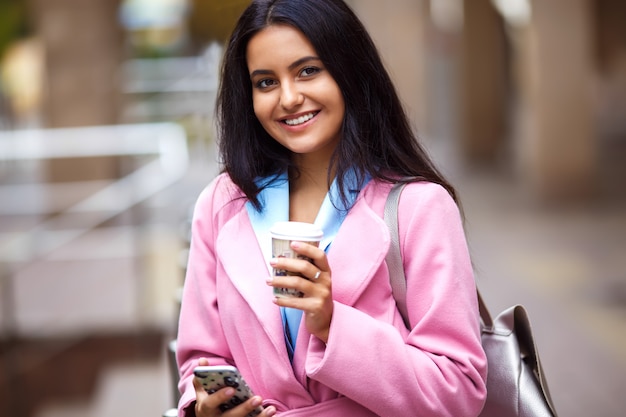 電話で話している美しい笑顔の美しい少女。ハンドバッグと一杯のコーヒーと電話を彼女の手で通りを歩いている若い美しいきれいな女の子