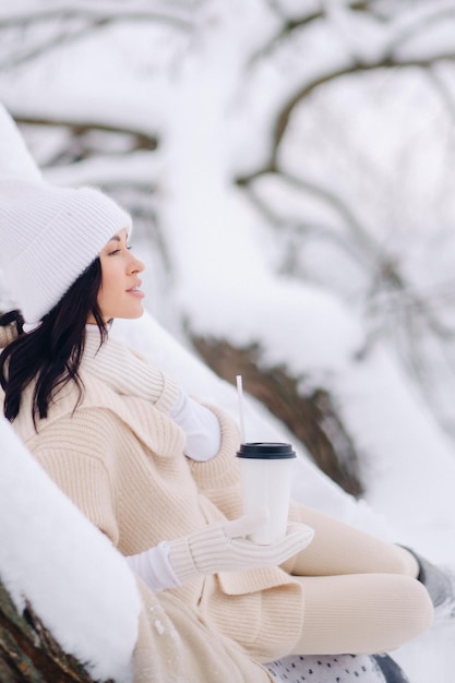 湖の近くの雪に覆われた冬の森でお茶を楽しんでいるベージュのカーディガンと白い帽子をかぶった美しい少女