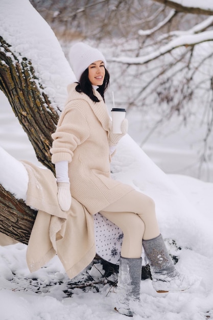 A beautiful girl with a beige cardigan and a white hat enjoying drinking tea in a snowy winter forest near a lake