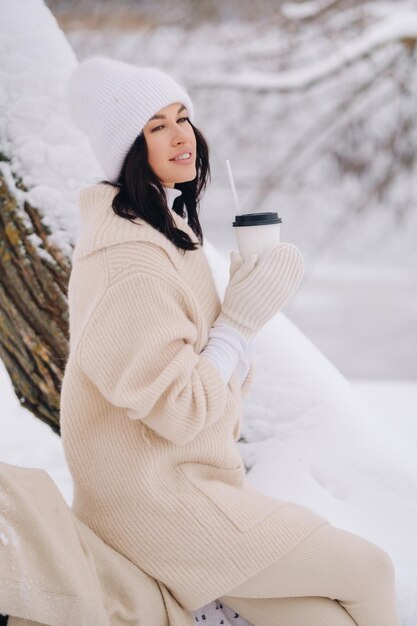 A beautiful girl with a beige cardigan and a white hat enjoying drinking tea in a snowy winter forest near a lake