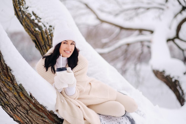 A beautiful girl with a beige cardigan and a white hat enjoying drinking tea in a snowy winter forest near a lake