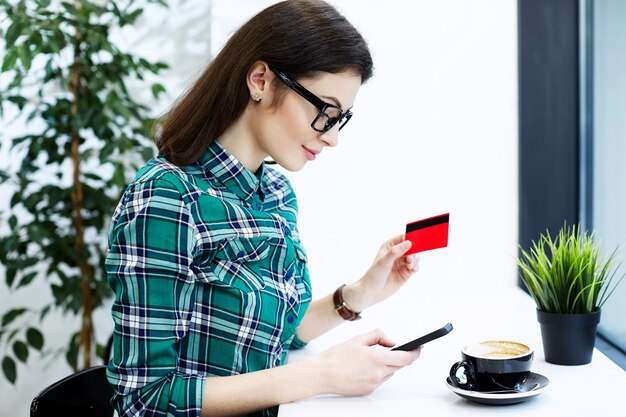 Beautiful girl with black hair wearing shirt and eyeglasses sitting in cafe with mobile phone and cup of coffee, holding credit card, online shopping.