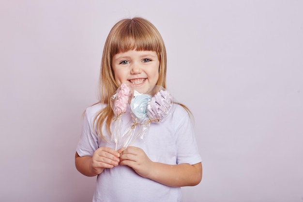 Beautiful girl with blond hair eats a Lollipop, round caramel on stick in hands of cheerful smiling girl. Baby girl with long hair in white t-shirt licks Lollipop. Russia, Sverdlovsk, 1 December 2018