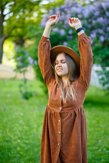 A beautiful girl with blond hair holds a hat in her hands and stands with her eyes closed near a lilac bush. Young woman in a garden with blooming trees