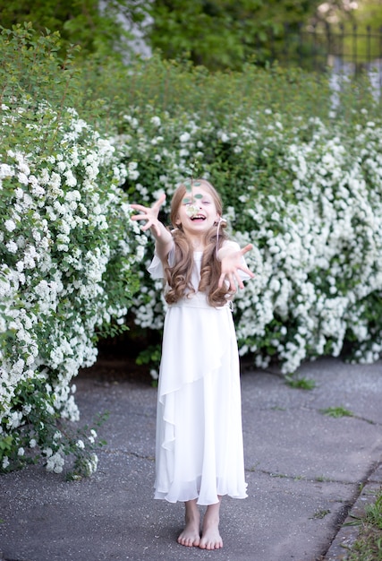 beautiful girl with blond long hair in a white delicate silk dress, having fun by blooming white spring flowers, throws white flowers in front of her, showing her hands