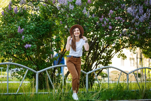 Beautiful girl with a brown hat standing near a lilac bush. Stylish young woman in a hat near a flowering tree. She laughs and enjoy life on a warm summer day.