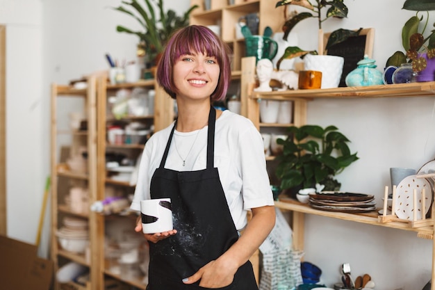 Beautiful girl with colorful hair in black apron and white T-shirt holding handmade mug in hand happily looking in camera spending time at pottery studio