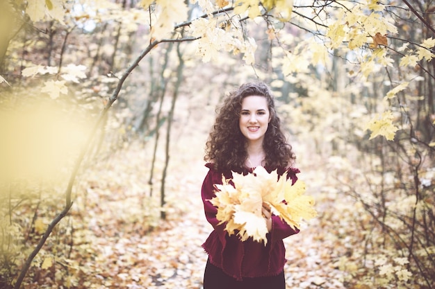 Beautiful girl with curly dark hair in a maroon top in an autumn park holding maple leaves and smili...