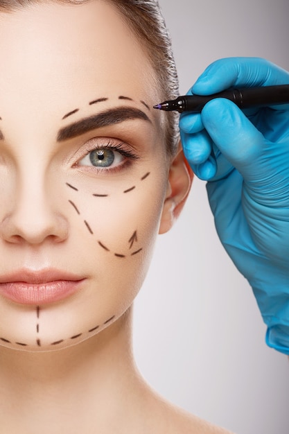 Beautiful girl with dark eyebrows at  wall, doctor's hands wearing blue gloves drawing perforation lines on face.
