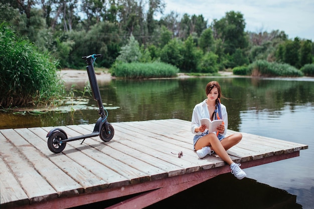 A beautiful girl with an electric scooter reads a book on a wooden bridge by the river. eco transport and health care concept.