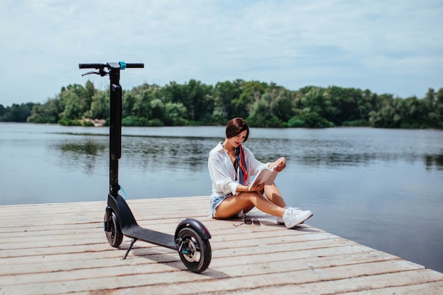 A beautiful girl with an electric scooter reads a book on a wooden bridge by the river. eco transport and health care concept.